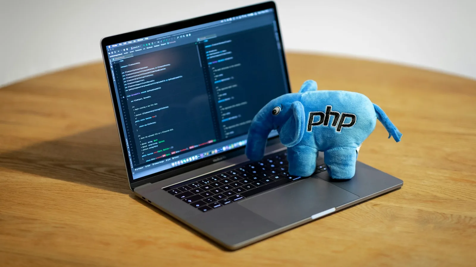 Blue plush toy of the PHP elephant mascot sitting on an open laptop showing lines of code