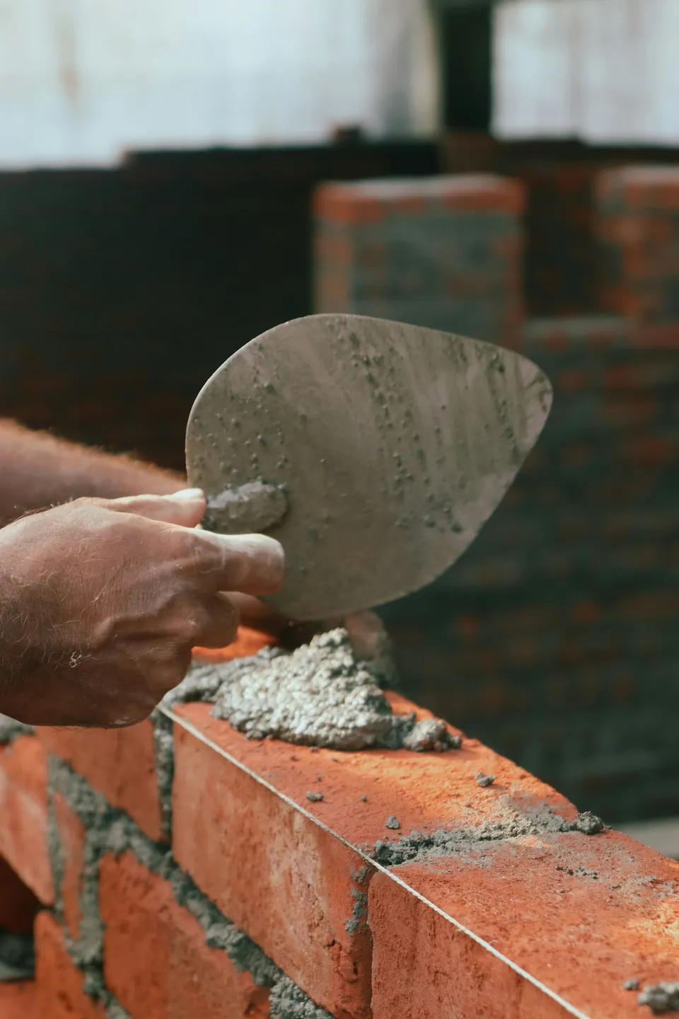Bricklayer's hand holding a trowel over a freshly mortared course of red bricks