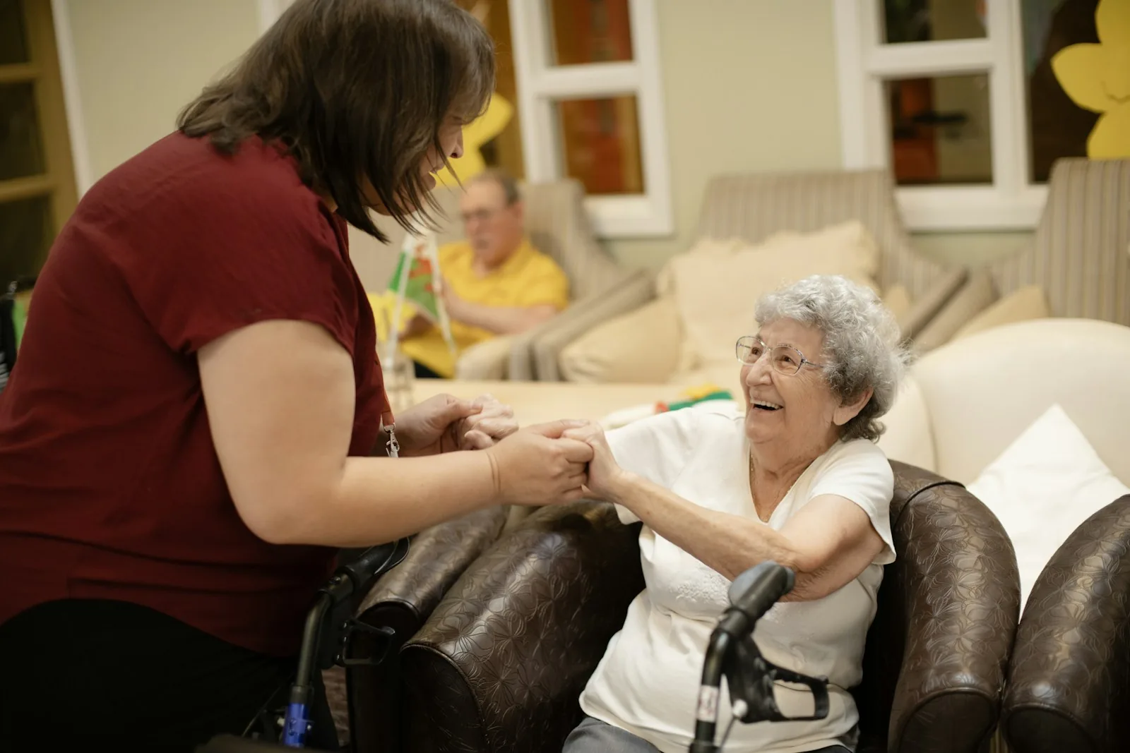 Carer holding the hands of a smiling older woman seated with her walking frame
