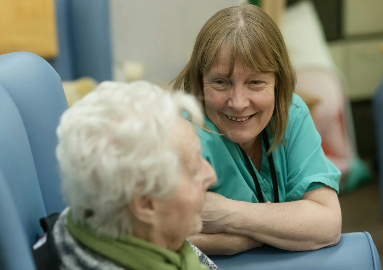 Carer in green scrubs leaning in to talk with an older woman sitting in a chair