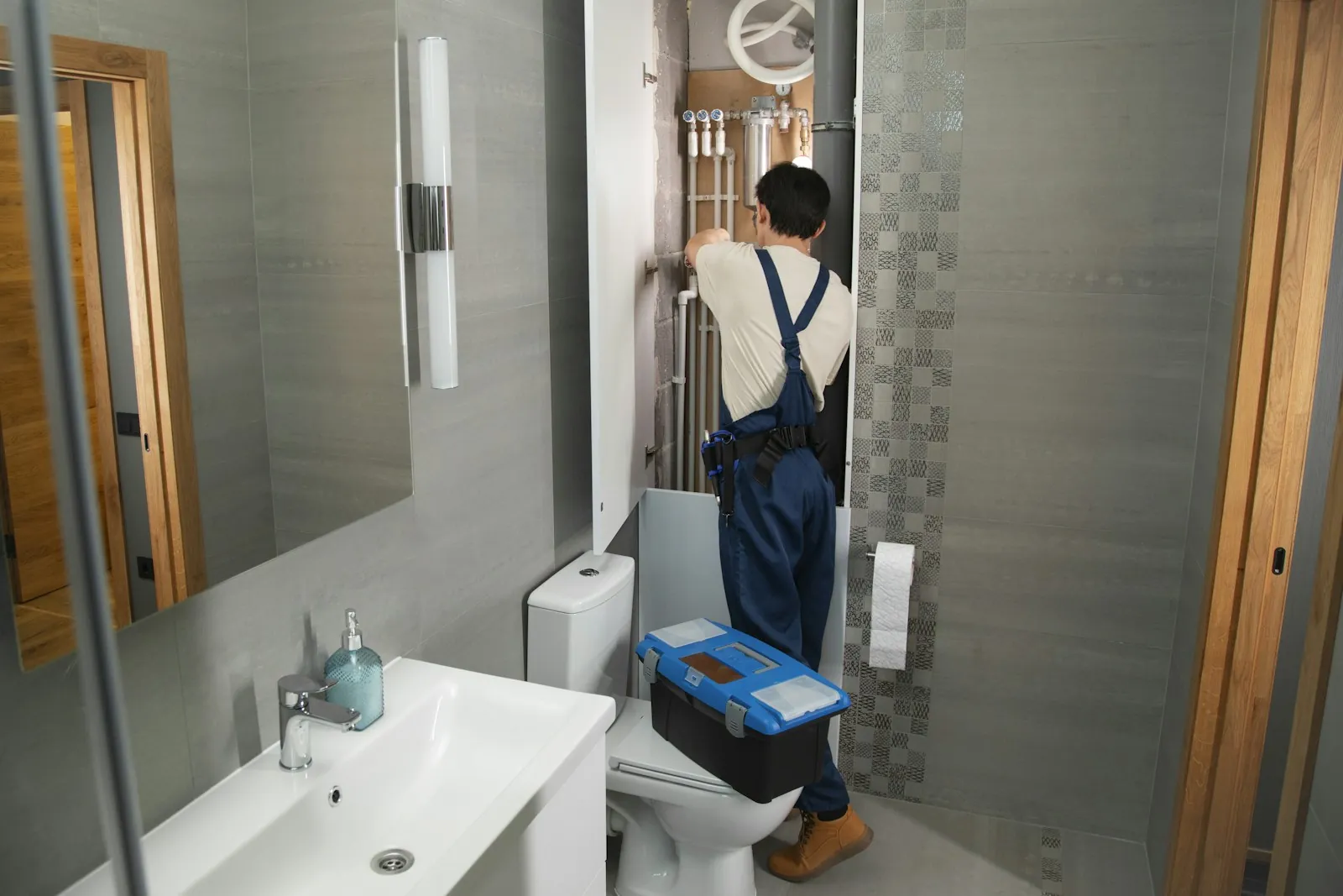 Plumber in navy overalls with a blue toolbox working in a modern tiled bathroom
